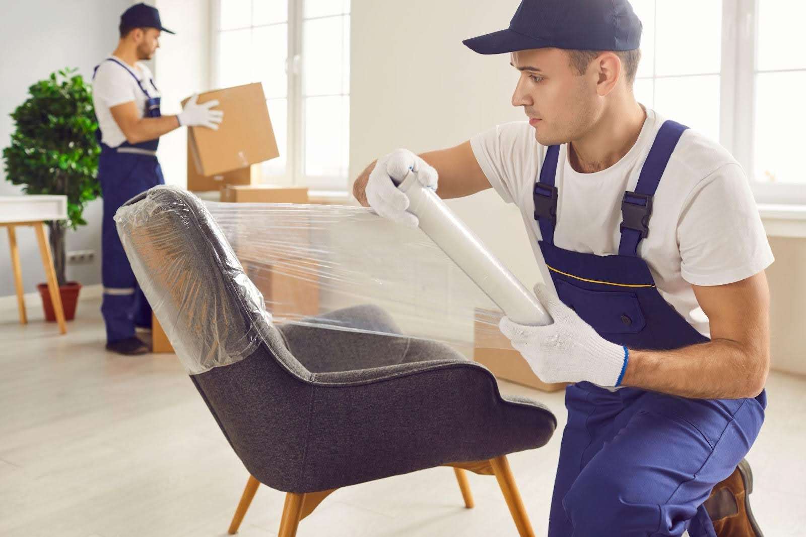 Mover with blue overalls and white gloves wraps a chair in plastic wrap while another Sturbridge MA mover moves boxes in the background.
