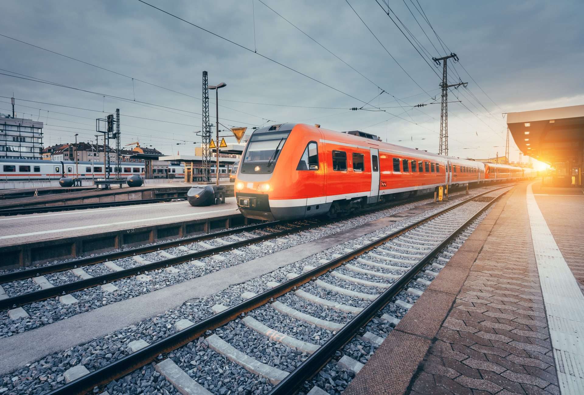 A modern red train sits at a railway station platform during sunset, with overhead power lines and city buildings in the background.